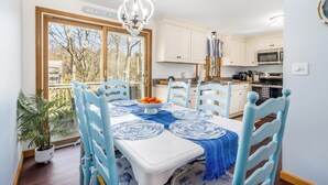 Een eethoek met een witte tafel, blauwe stoelen en een schaal met fruit. Er is een groot raam met uitzicht op bomen en een bordje met de tekst 