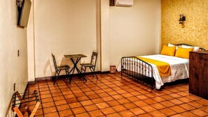 A small, compact room with a tiled floor, a bed with yellow bedding, a small table with chairs, and a wall-mounted TV. - Hotel Boutique Casa de las Flores     (Huajuapan)