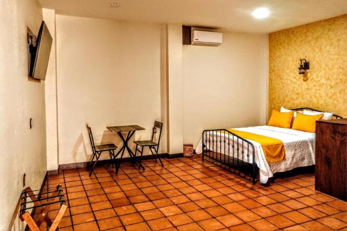 Une petite pièce, de taille modeste, avec un sol carrelé, un lit recouvert d’une literie jaune, une petite table avec des chaises et un téléviseur fixé au mur.