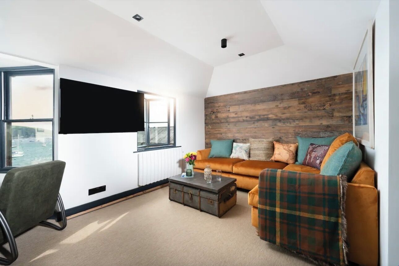 A modern living room with a large window, a flat-screen TV, a wooden accent wall, a sofa with a plaid throw, and a coffee table.