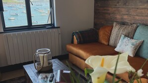 A cozy living room with a view of boats through a large window, a wooden wall, a brown sofa with cushions, and a coffee table with a candle, book, and flowers. - Captains Loft (Falmouth)