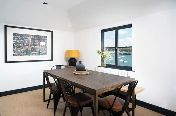 A dining area with a wooden table and chairs, a framed artwork on the wall, a lamp, and a view of the sea through a window. - Captains Loft (Falmouth)