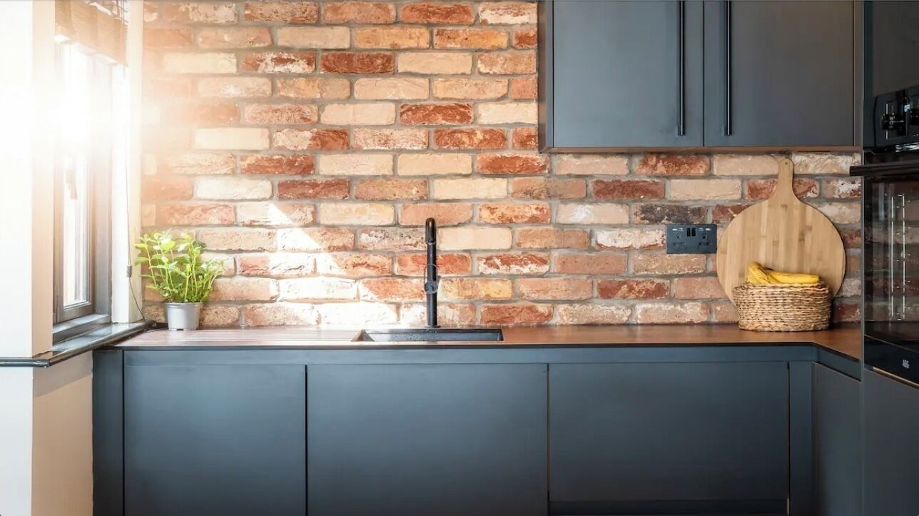A kitchen with dark cabinets, a brick wall, a wooden cutting board, and a basket of bananas.