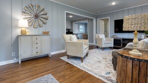A living room with a large decorative wall piece, a wooden cabinet, two armchairs, a round coffee table, and a television. - Cozy & Tasteful 3Br Cottage Walkable to Downtown (Franklin)