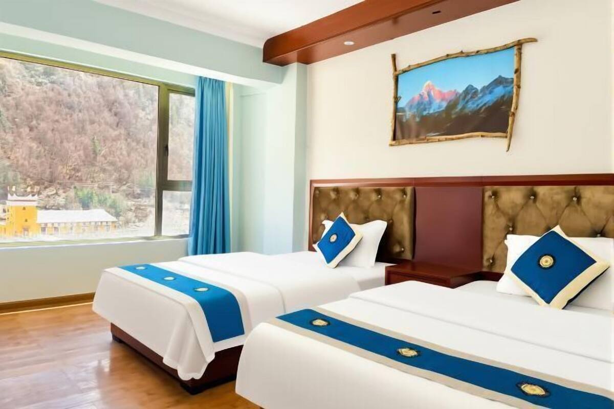 Une chambre d’hôtel avec deux lits, une grande fenêtre donnant sur des arbres et un tableau représentant une montagne accroché au mur.