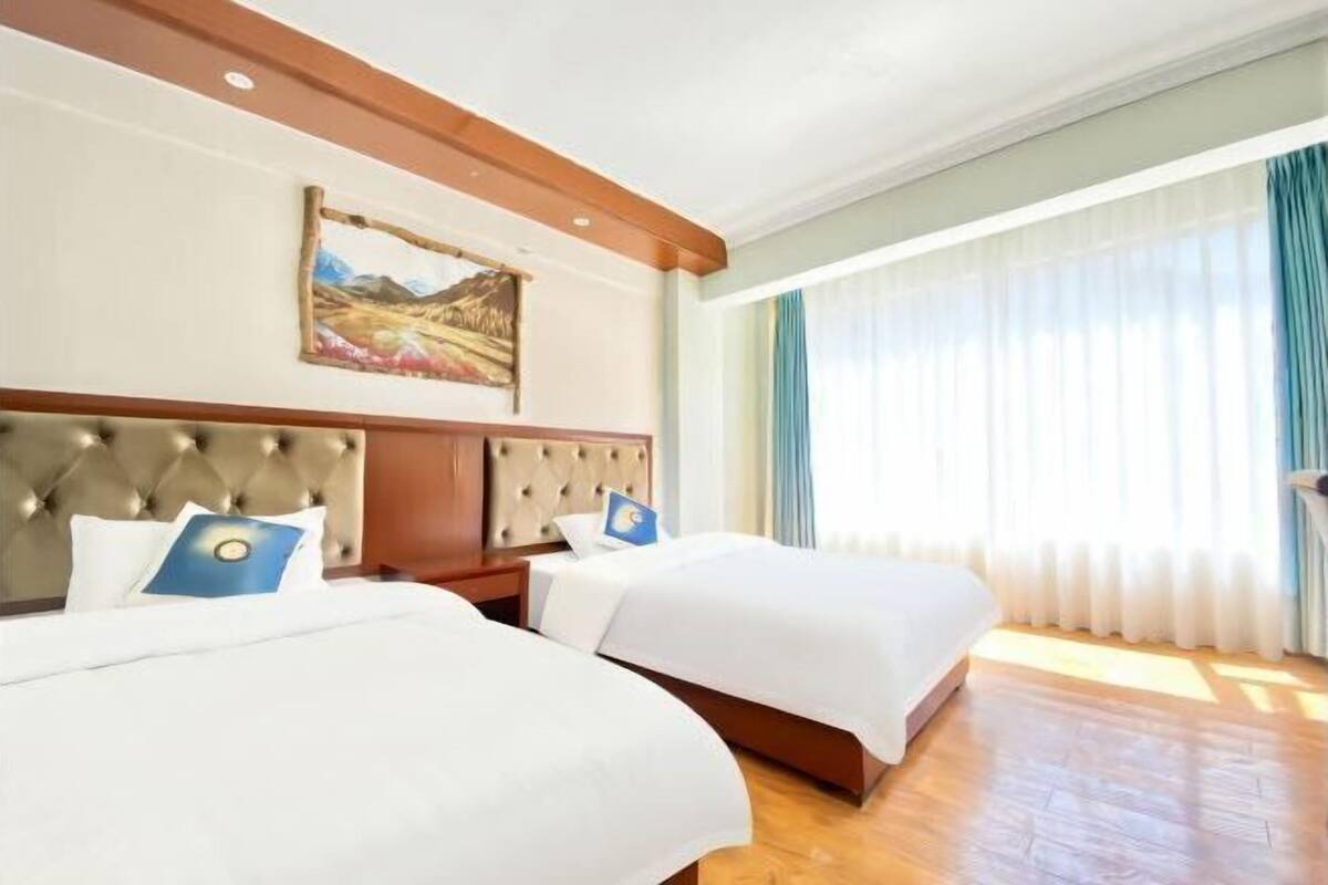 Une chambre d’hôtel avec deux lits, une tête de lit capitonnée, un tableau au mur et une grande fenêtre avec des rideaux.