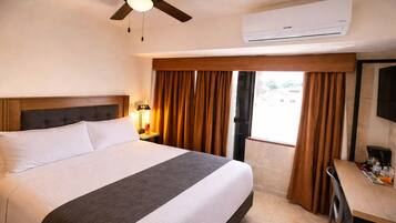 A hotel room with a bed, a desk, a ceiling fan, an air conditioner, and a window with curtains.
