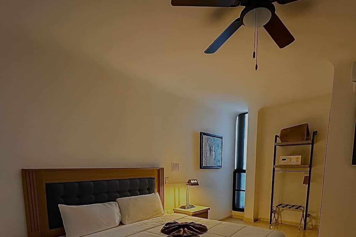 Une chambre d’hôtel avec un grand lit, un ventilateur de plafond, une table de chevet, une lampe et une petite étagère avec une radio.