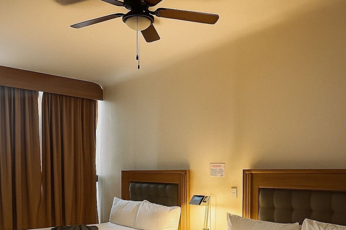 Une chambre d’hôtel avec deux lits, un ventilateur de plafond, une table de chevet avec une lampe et une fenêtre avec des rideaux.