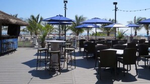 Un espace extérieur aménagé en terrasse, avec des tables et des chaises, des parasols bleus et une vue sur l’océan.
