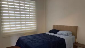 Een slaapkamer met een bed, nachtkastje en een raam met jaloezieën.