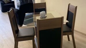 Een moderne eethoek met een glazen tafel, houten stoelen, een zwarte bank en een groot raam met jaloezieën.