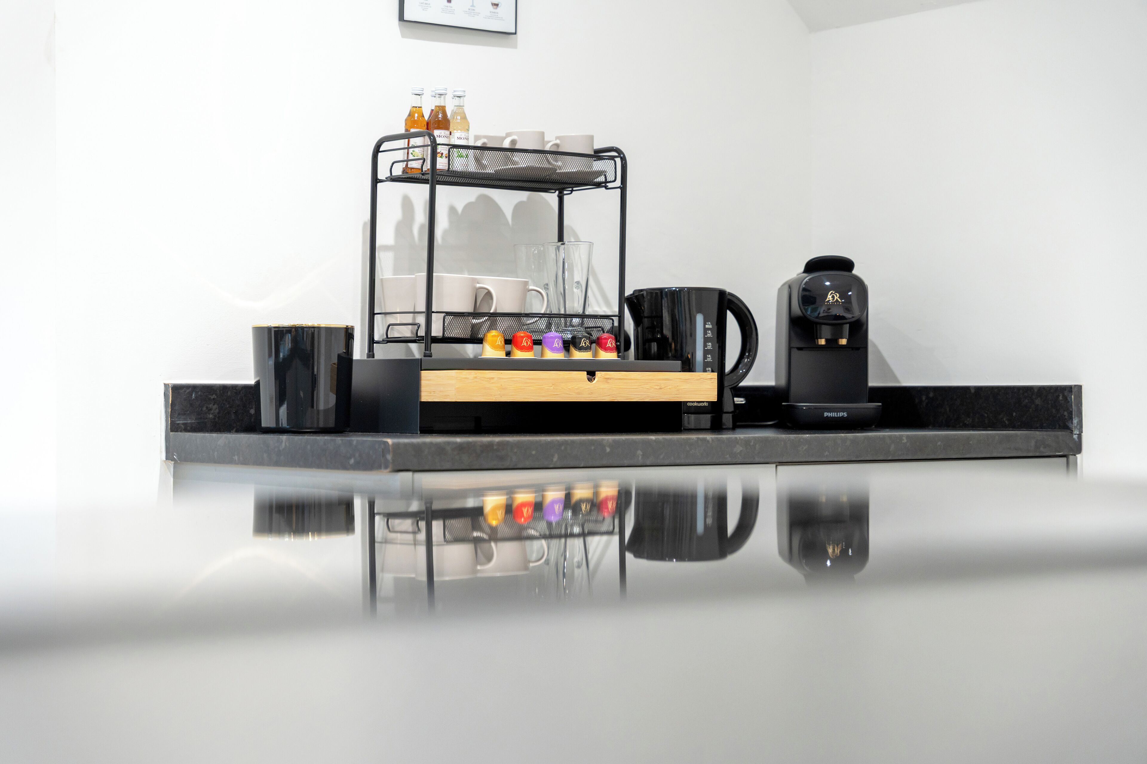 A clean, modern coffee station with a coffee maker, a tray with cups and bottles, and a coffee pot on a reflective countertop.