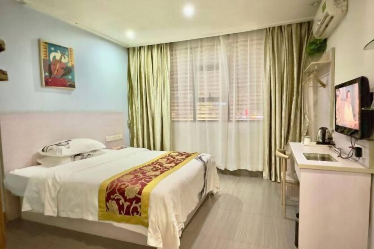 A hotel room with a bed, a desk with a sink, a television, and a window with curtains.