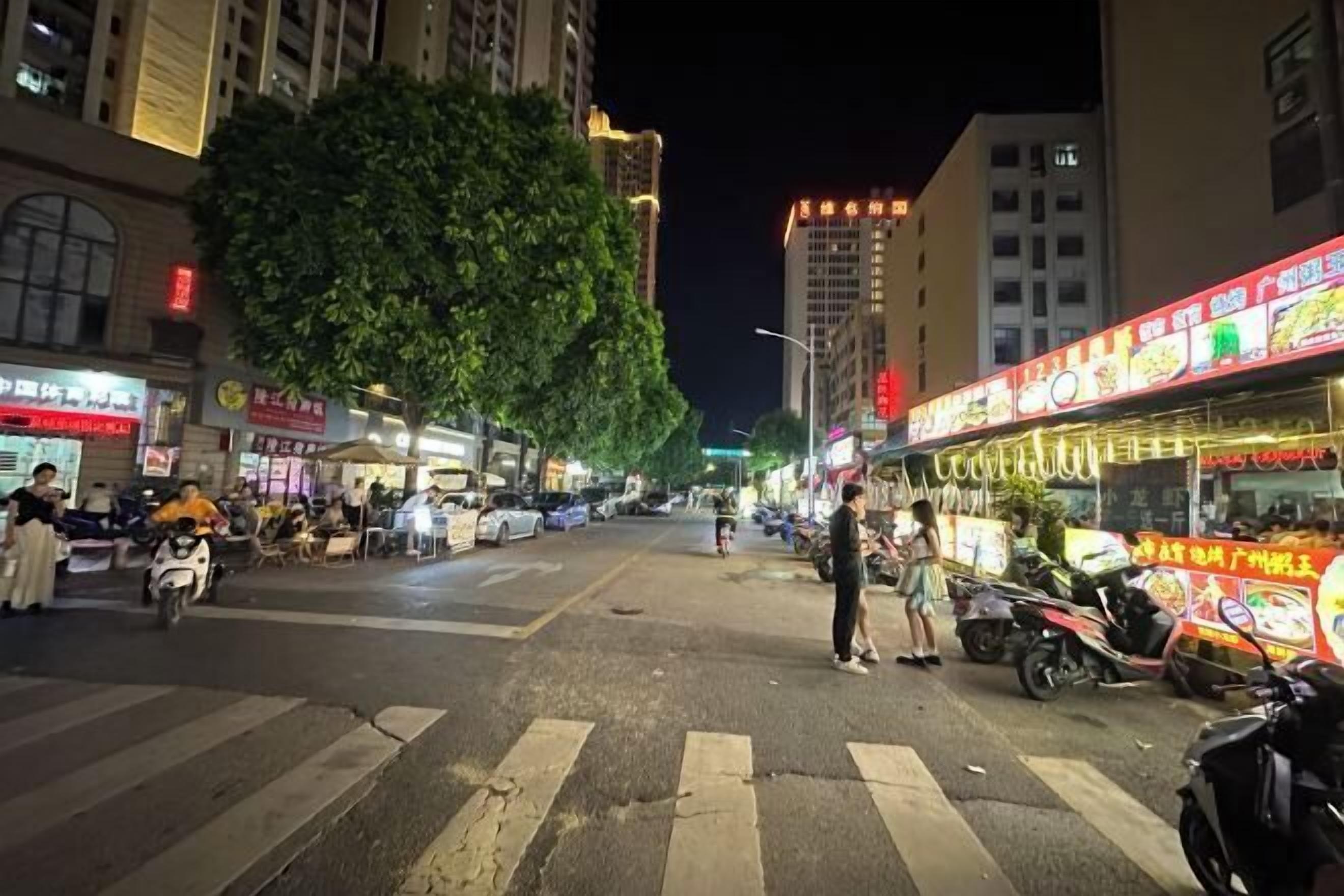 Escena nocturna en la calle con un paso de peatones, motocicletas estacionadas y un puesto de comida con mucha luz.