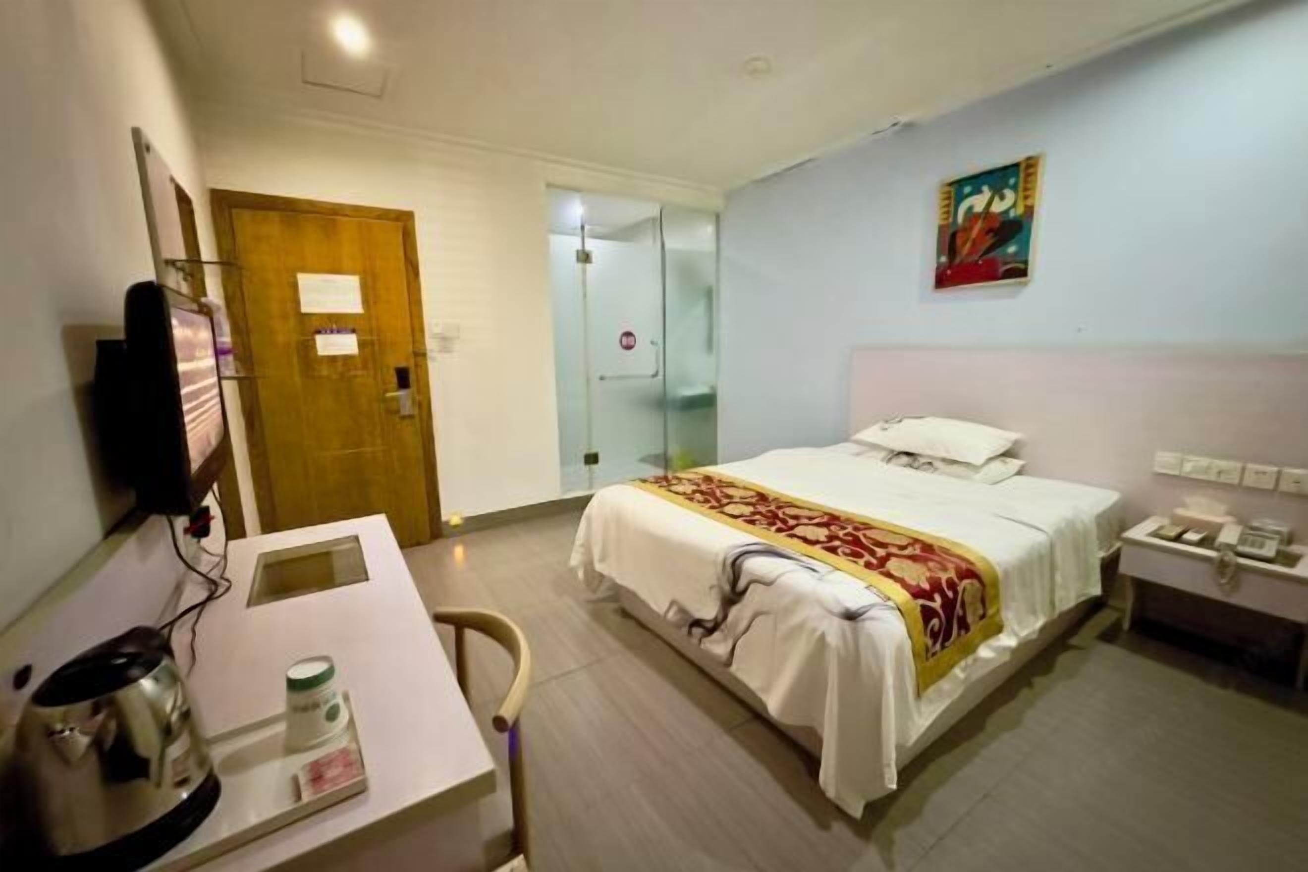 Une chambre d’hôtel avec un lit, une table de chevet, un bureau avec une bouilloire, une chaise et un tableau au mur.