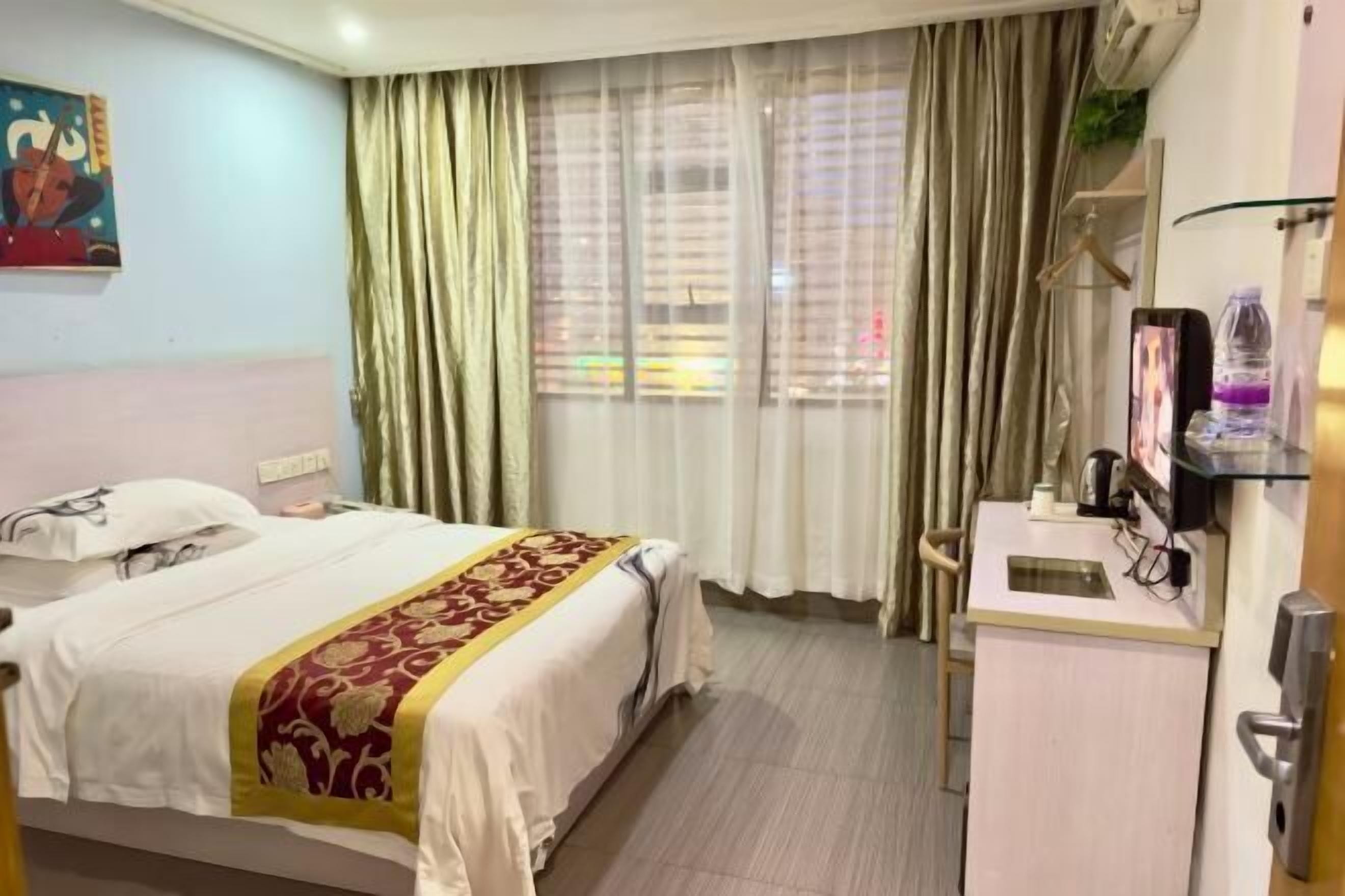 A hotel room with a bed, a desk, a window with curtains, and a painting on the wall.