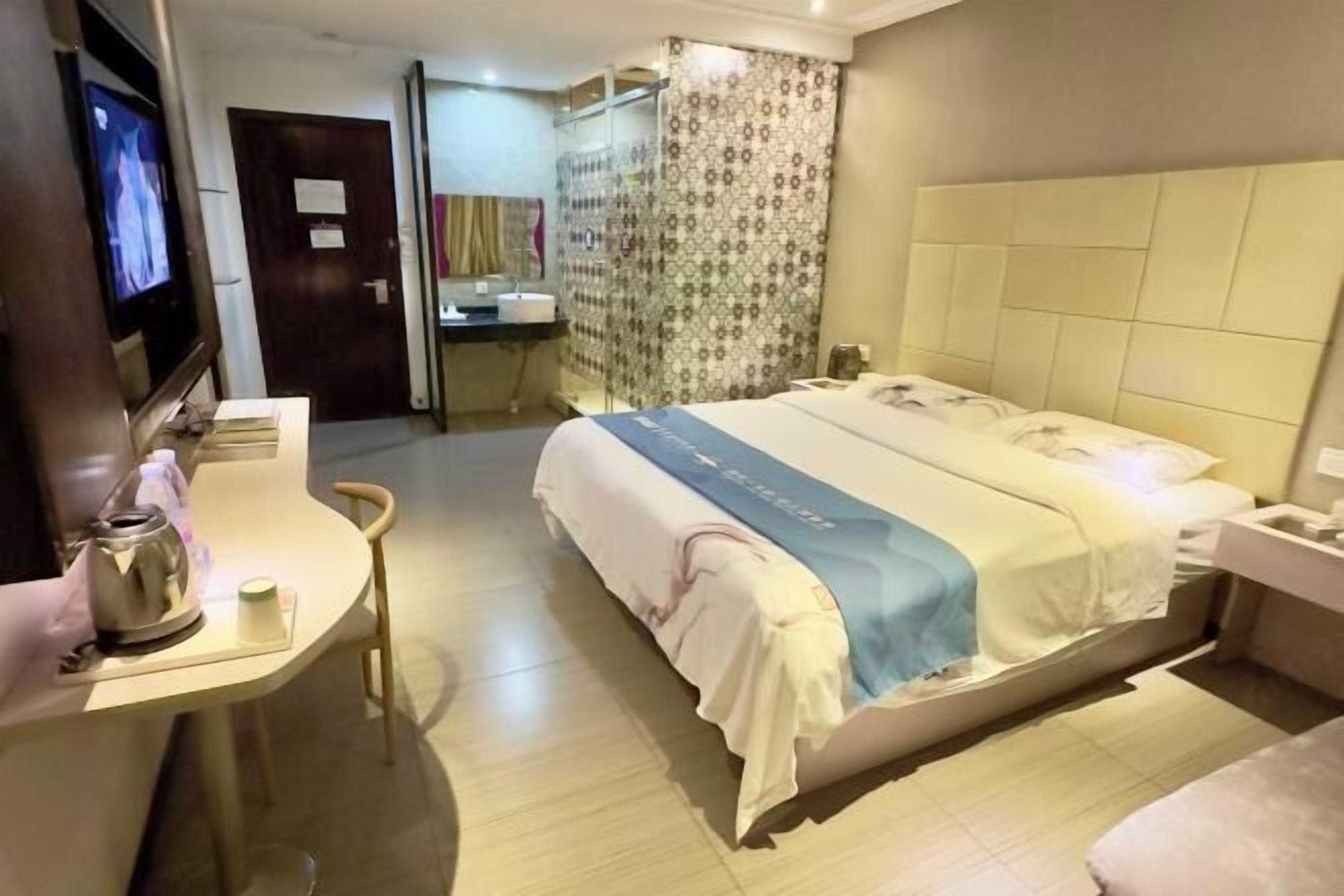 A hotel room with a bed, a desk with a kettle, a television, and a bathroom visible through a glass door.
