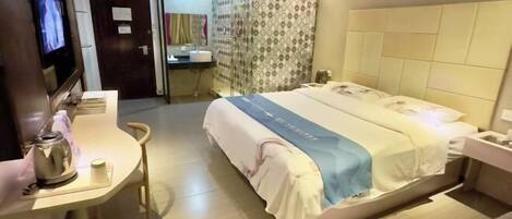A hotel room with a bed, a desk with a kettle, a television, and a bathroom visible through a glass door.
