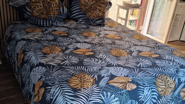 A bedroom with a bed covered in a tropical leaf-patterned bedspread, wooden slats on the wall, and a view of the outside through a window.