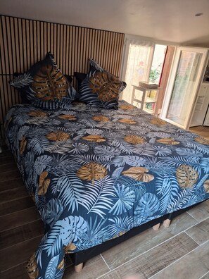 A bedroom with a bed covered in a tropical leaf-patterned bedspread, wooden slats on the wall, and a view of the outside through a window.