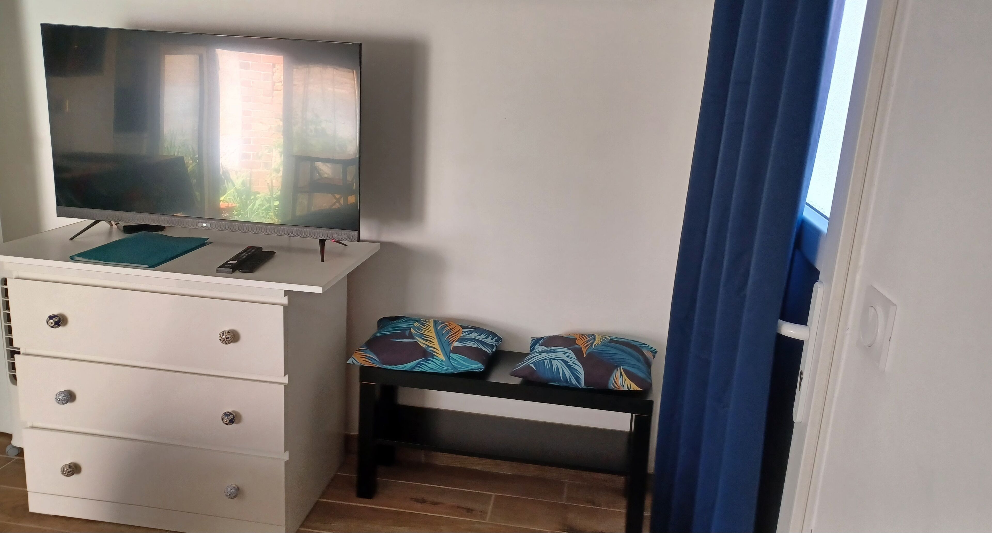 A room with a TV on a white dresser, a bench with patterned cushions, and a blue curtain.