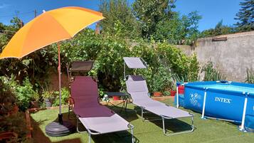 A backyard with two sun loungers, a large yellow umbrella, a blue inflatable pool, and various potted plants.