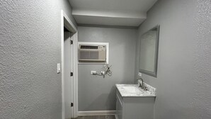 Une petite salle de bain avec un lavabo blanc, un miroir et une fenêtre équipée d’un appareil de climatisation.