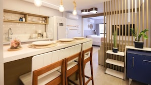 A modern kitchen with white cabinetry, a built-in oven, and a coffee machine. There are wooden chairs and a blue cabinet with a handle. - Studio Shopping OKA Floripa - Campeche (Florianopolis)