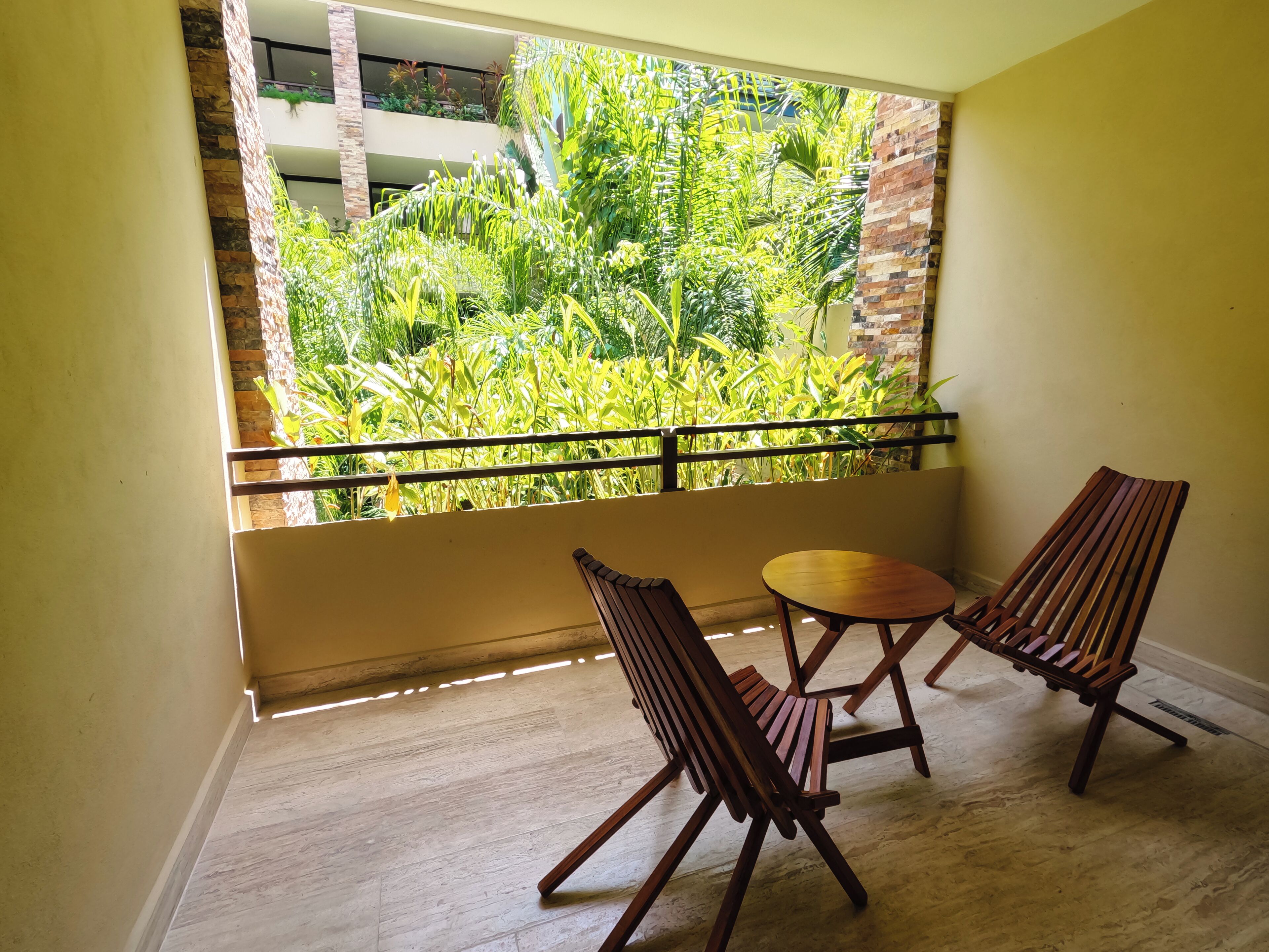 A balcony with two wooden chairs and a small round table, overlooking a lush garden with various plants and a building in the background.