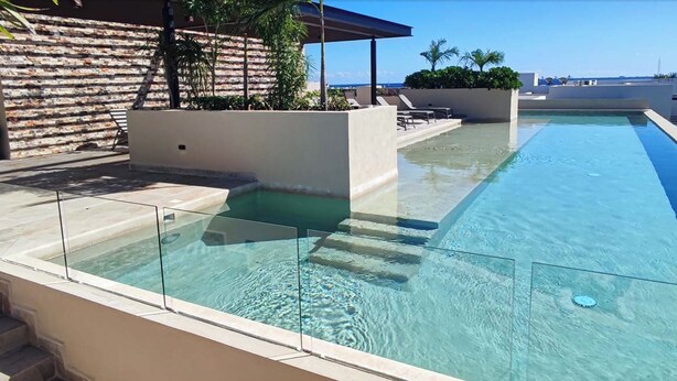 Ein klarer, rechteckiger Pool mit Glasbarriere, umgeben von einer niedrigen Steinmauer und einem überdachten Sitzbereich mit Liegestühlen.