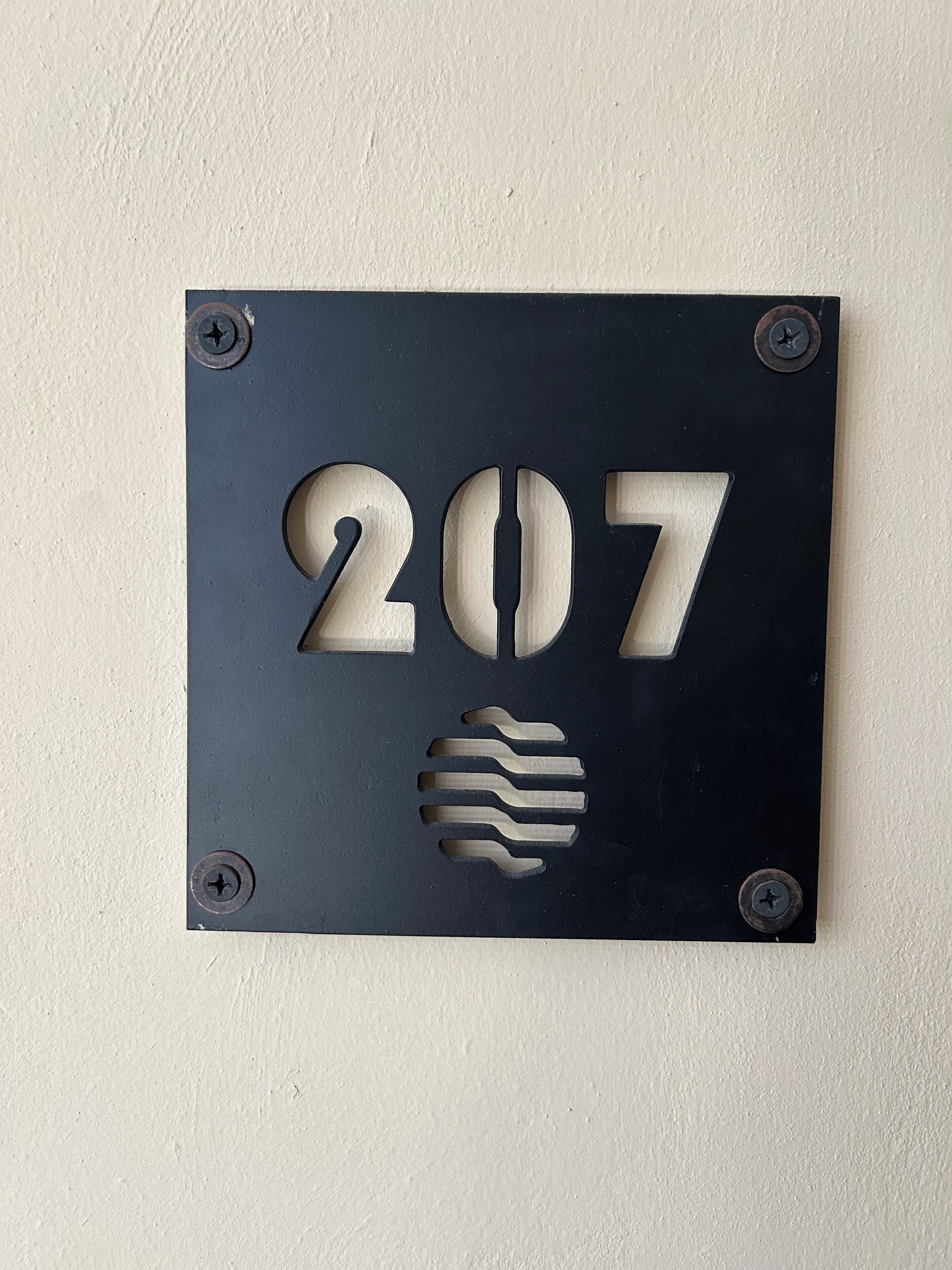 A metal number plate with the number 207, mounted on a wall with four screws.