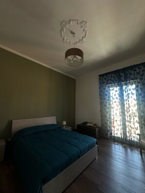 Een slaapkamer met een bed, nachtkastje, lamp, raam met vlindergordijnen en een plafondlamp.