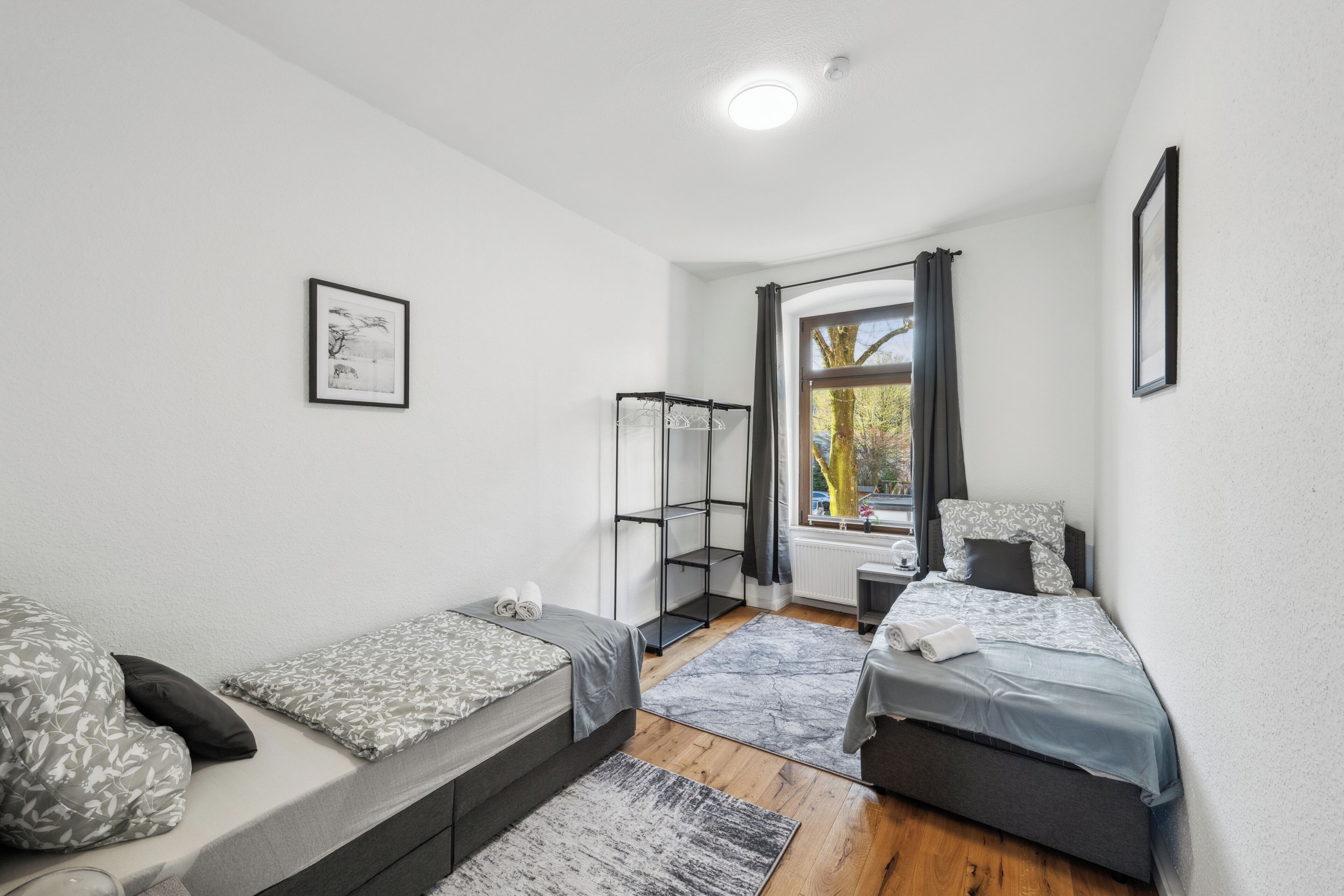 Un dormitorio pequeño y moderno con dos camas individuales, una ventana con cortinas, un radiador y un cuadro enmarcado en la pared.