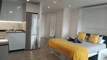 A modern hotel room with a bed, bedside table, and a kitchenette area.