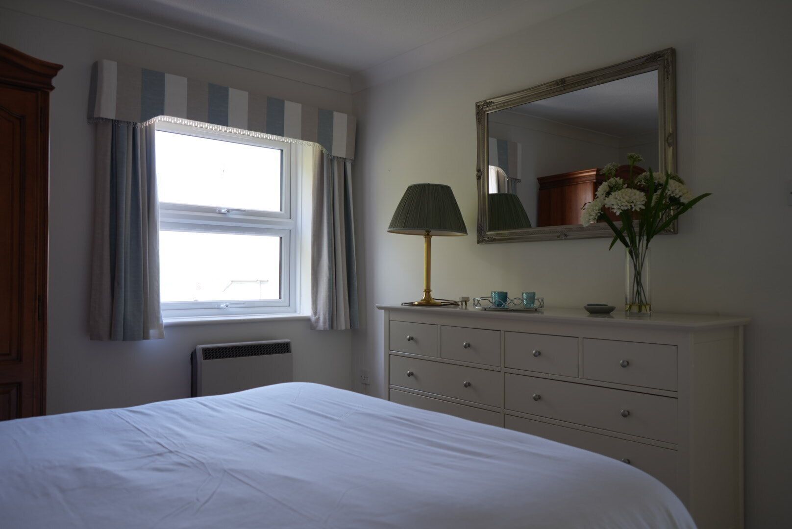 A bedroom with a bed, a dresser, a mirror, a window with curtains, and a lamp.