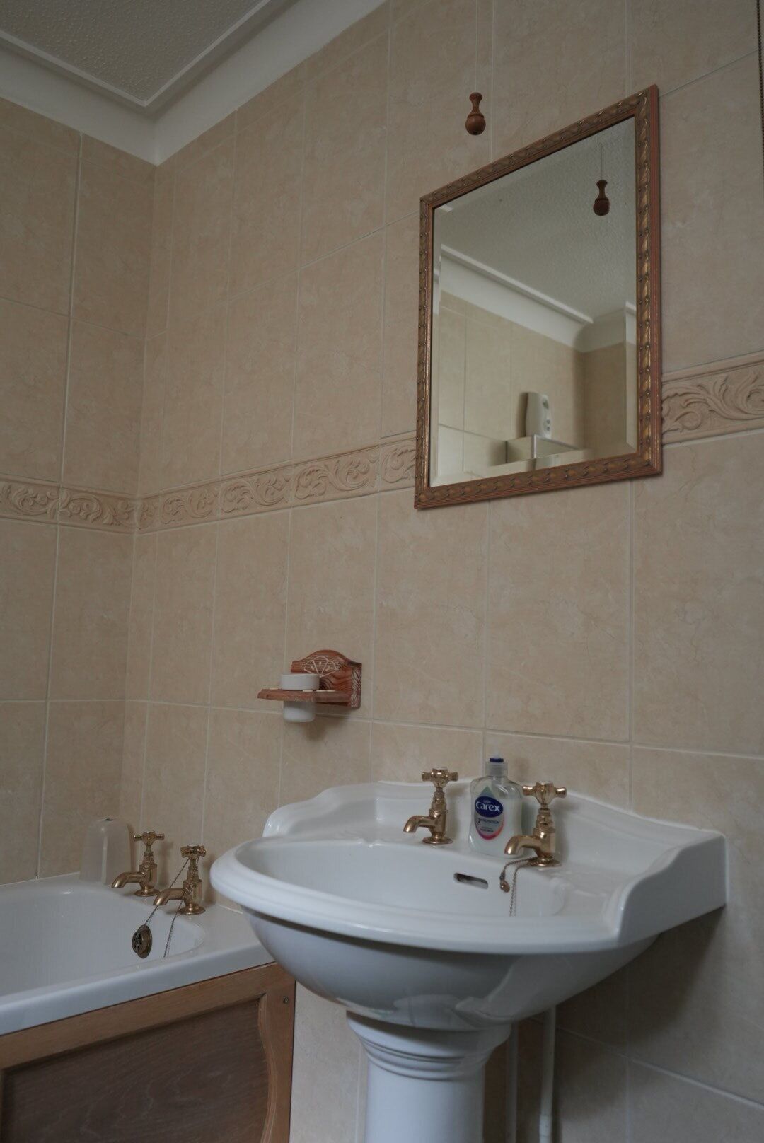 A bathroom with a white pedestal sink, two gold faucets, and a mirror with a wooden frame.