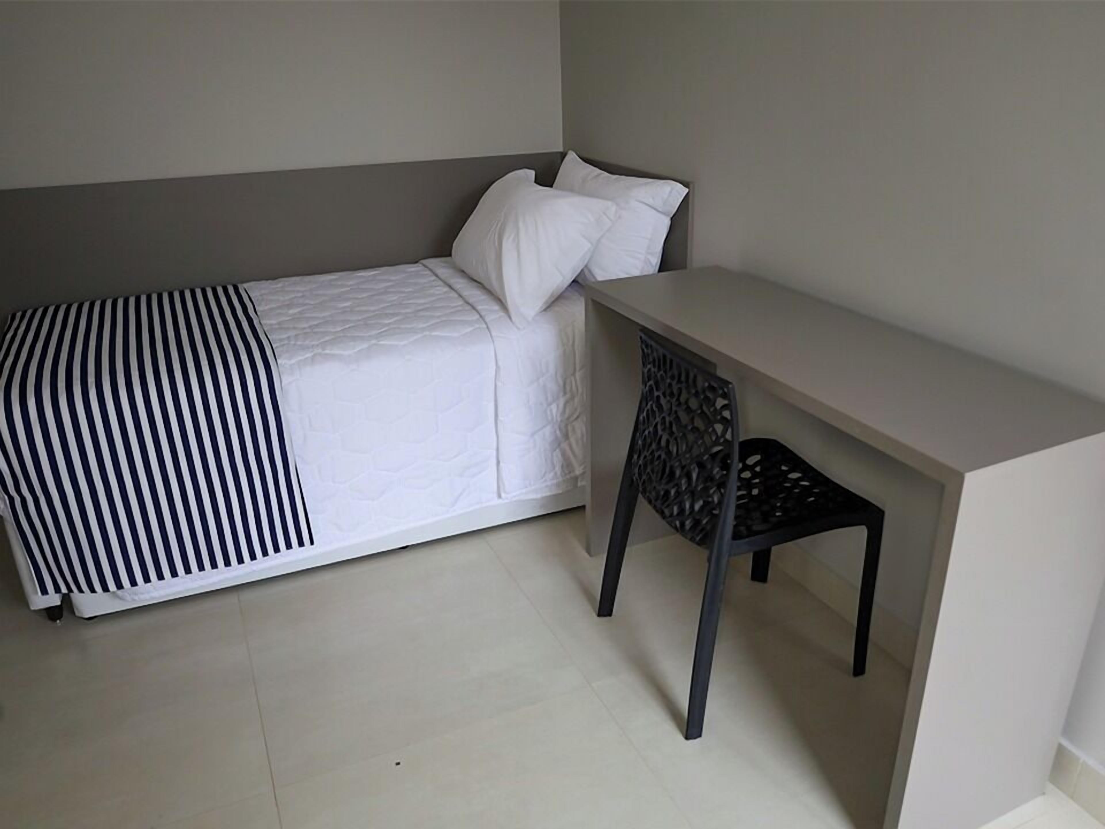A single bed with a striped blanket, a white pillow, and a black chair under a desk in a room with plain walls and a tiled floor.