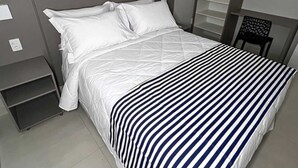 Una cama bien hecha con ropa blanca y una manta a rayas, una mesita de noche pequeña y una silla con un diseño original en el respaldo.