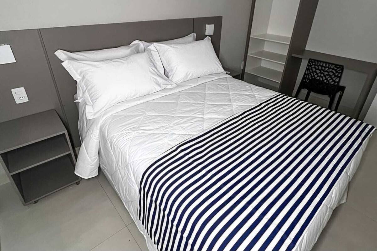 A neatly made bed with white bedding and a striped blanket, a small bedside table, and a chair with a unique backrest design.