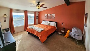 Un dormitorio con cama, ventilador de techo, ventana con cortinas, mesita de noche, lámpara y maleta.