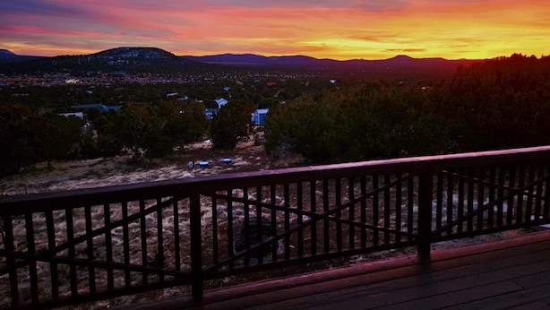 Vista del atardecer desde un balcón con barandilla de madera, con vista a un pueblo y montañas distantes.