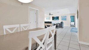 A spacious dining area with a long table and white chairs, leading to a kitchen area with a dining table and chairs, and a living area with a TV and artwork on the wall.