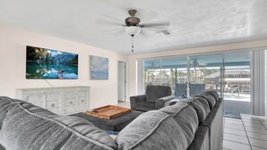 A spacious living room with a gray sectional sofa, a wooden coffee table, and a ceiling fan. There are two paintings on the wall and a large window offering a view of a pool and outdoor furniture.