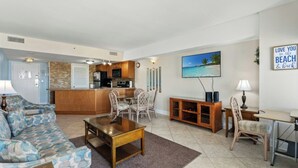 A living room with a sofa, coffee table, and dining area with chairs. There is a kitchen area with a refrigerator and microwave. A painting of a beach is on the wall. - Ocean View Palace Resort 1813- 2BR- Pool, Hot Tub (Myrtle Beach)