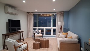 A modern living room with a TV, sofa, armchair, coffee table, and a large window with curtains.