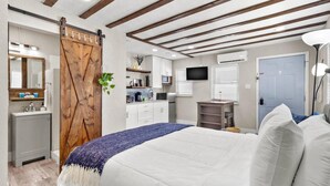 Un dormitorio con cama, puerta de madera, televisor, escritorio y baño con lavamanos y espejo.