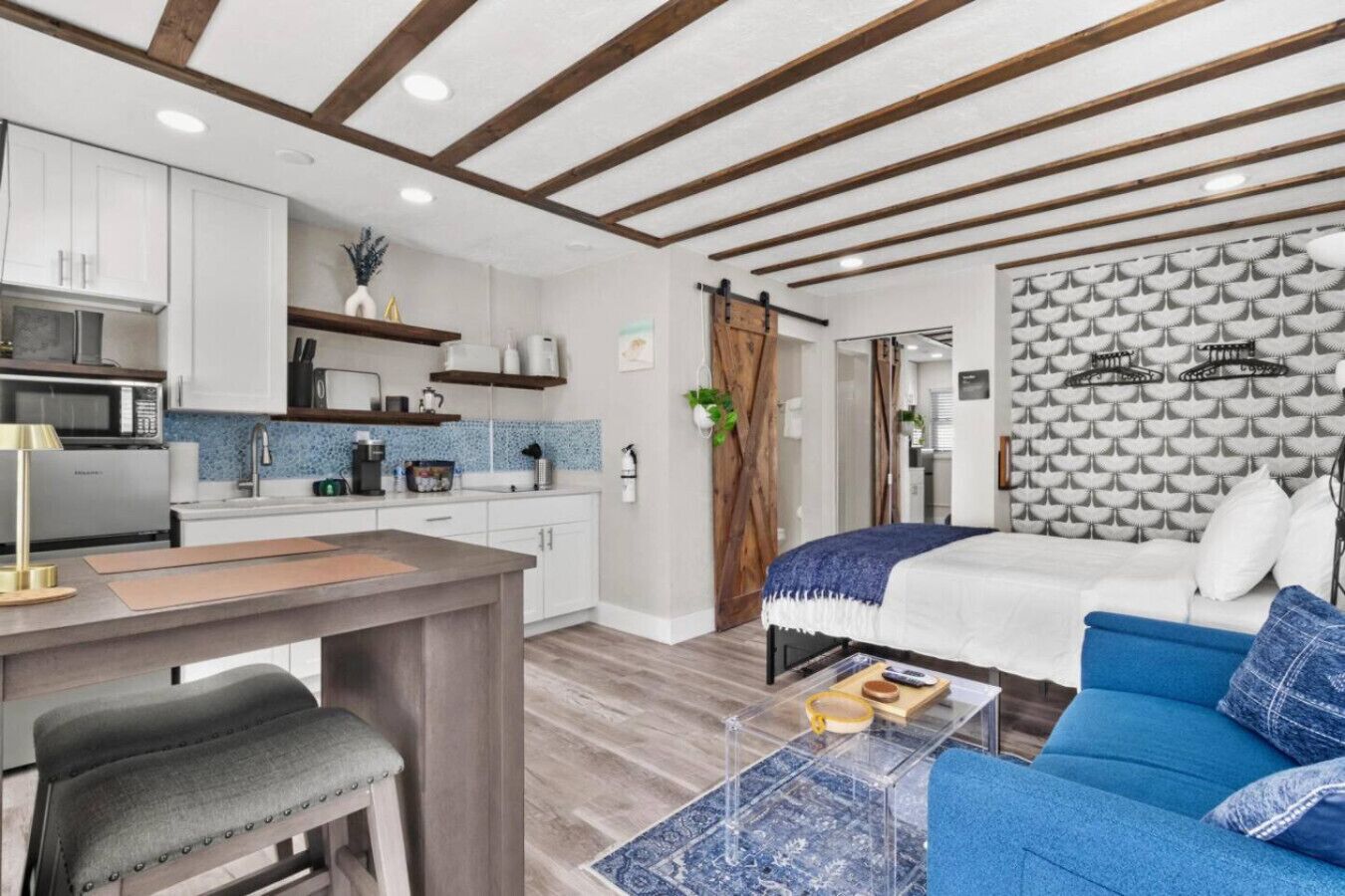 Une cuisine moderne avec des meubles blancs, un four à micro-ondes et une machine à café. On y trouve un coin repas avec un banc et une table. La pièce est équipée d’un lit avec une couverture et des oreillers bleus, et d’un mur décoré d’un motif.