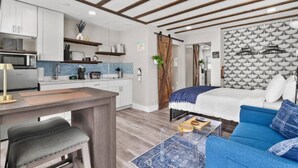 Eine moderne Küche mit weißen Schränken, einer Mikrowelle und einer Kaffeemaschine. Es gibt eine Essecke mit einer Bank und einem Tisch. Der Raum verfügt über ein Bett mit blauer Decke und Kissen sowie eine Wand mit Musterung.