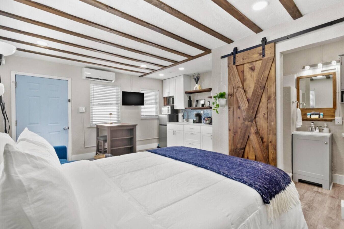 A bedroom with a bed, a desk, a TV, a wardrobe, and a bathroom with a mirror and sink.